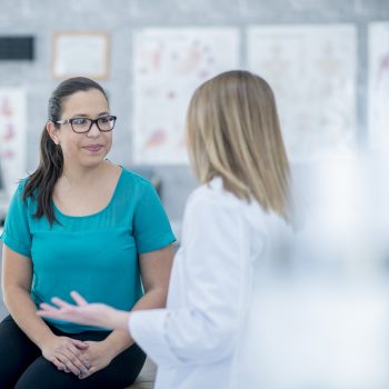 A doctor has her back to the camera while talking to an older female patient. The patient is smiling at her while her hands rest in her lap.